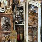 A rustic vintage-style decorative cabinet or wall display filled with antique glass bottles, jars, a hanging lantern, and distressed wood panels. The scene is tightly framed and appears to show the product as a styled decor arrangement rather than a clean isolated studio shot.