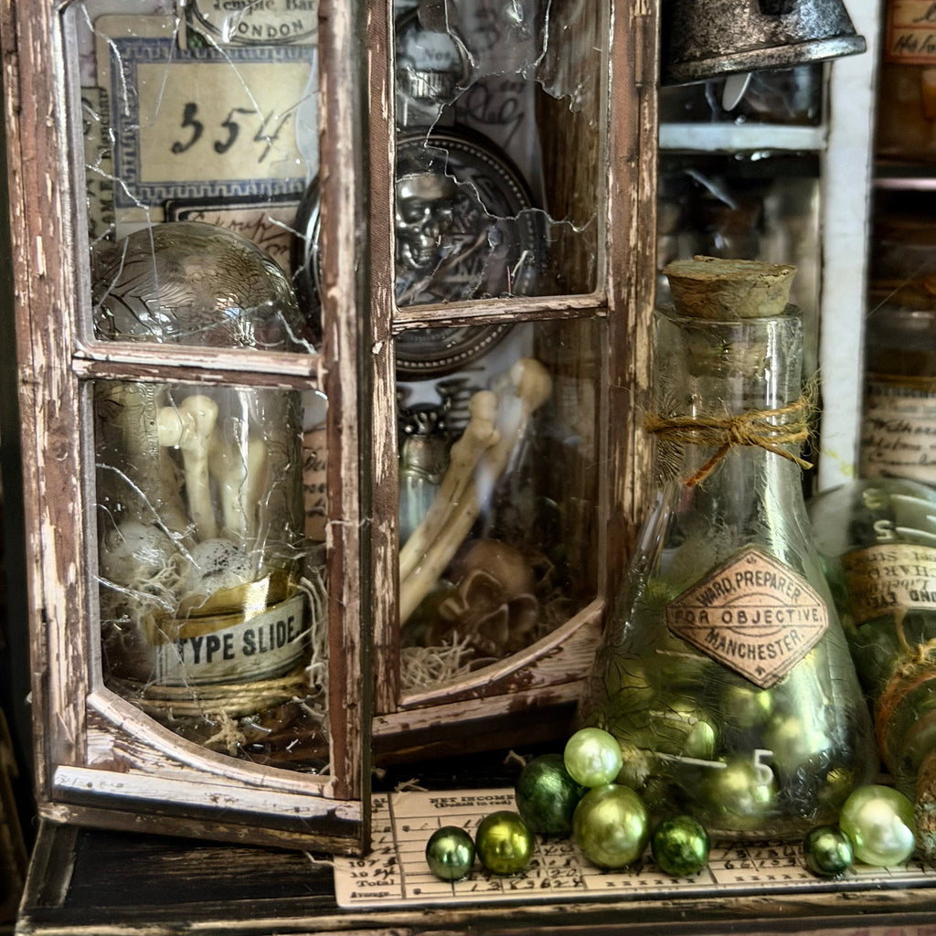 A vintage-style decorative lantern and glass bottle arrangement with bones, metallic ornaments, aged paper labels, and distressed wood framing on a dark tabletop.