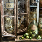 A vintage-style decorative lantern and glass bottle arrangement with bones, metallic ornaments, aged paper labels, and distressed wood framing on a dark tabletop.