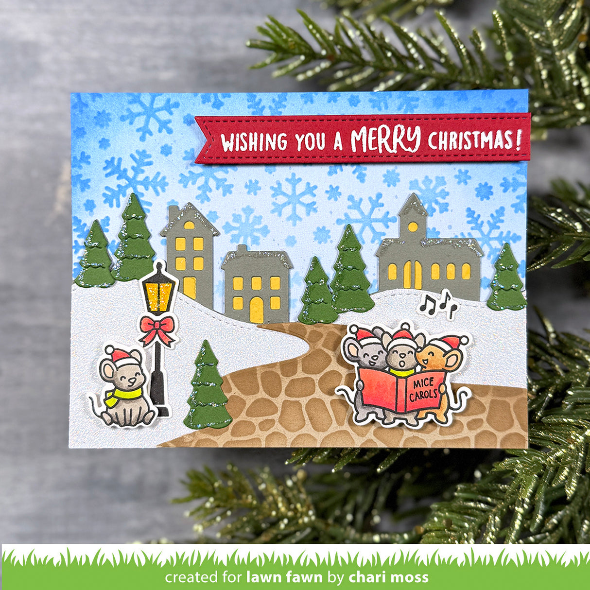 Christmas card with snowy village scene and mice reading a book, surrounded by festive greenery.