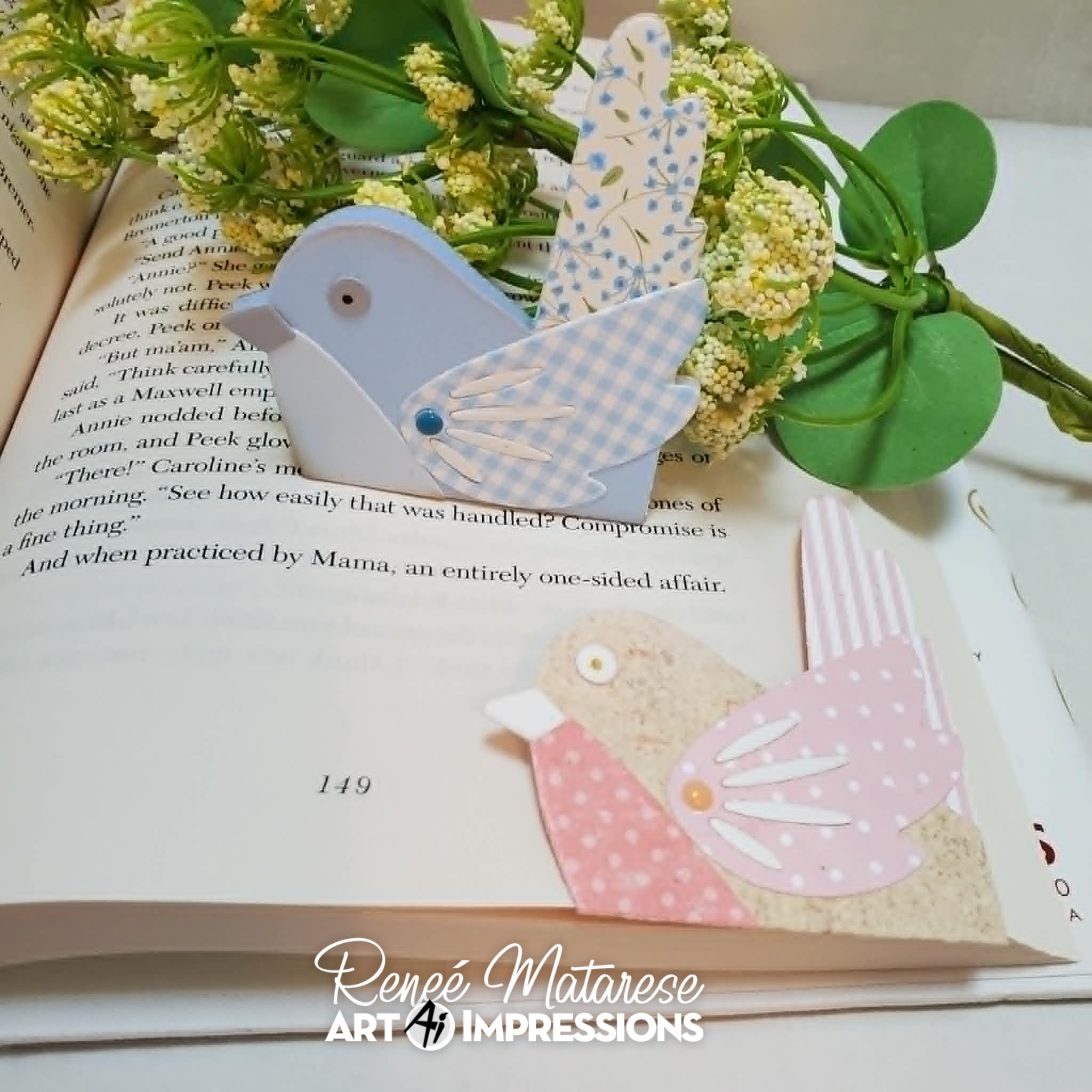 Two handmade bird-shaped bookmarks made from light blue, white patterned, and pink patterned cardstock with white paper cutouts, displayed on an open book with green leaves and white flowers.